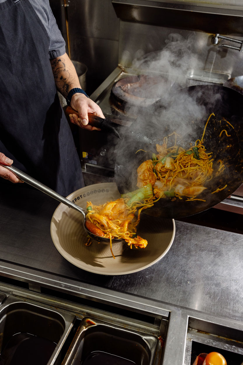 Chef cooking hotpot ingredients in a wok with steam rising.